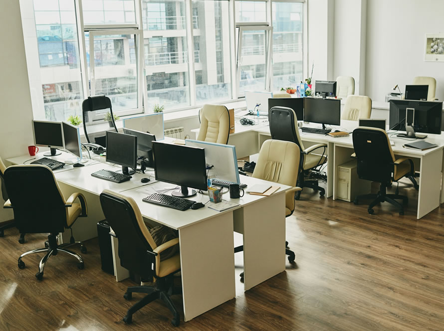 Espacio de trabajo compartido con escritorios blancos, equipos informáticos y grandes ventanales.